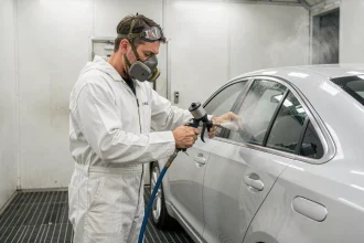 Technicien carrossier appliquant du vernis transparent au pistolet à peinture sur un panneau de carrosserie argent dans une cabine de peinture professionnelle